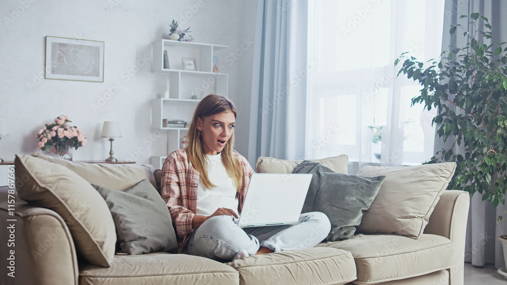 Young woman sitting on a comfortable sofa in her living room, working from home on her laptop, looking amazed by some good news she's receiving Young woman sitting on a comfortable sofa in her living room, working from home on her laptop, looking amazed by some good news she's receiving