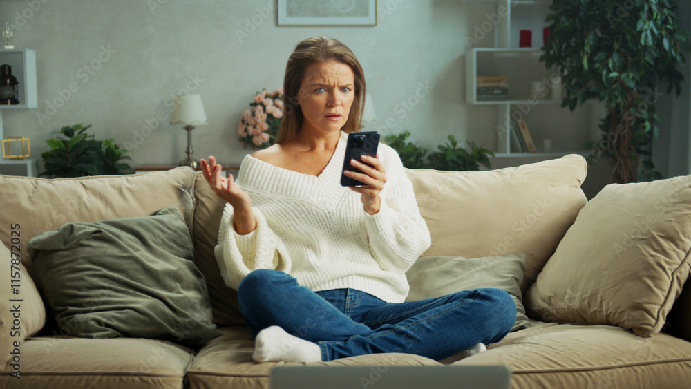 Obraz premium Middle aged woman sitting on a cozy sofa at home, looking confused while reading a message on her smartphone, gesturing with her hand, and displaying a range of negative emotions