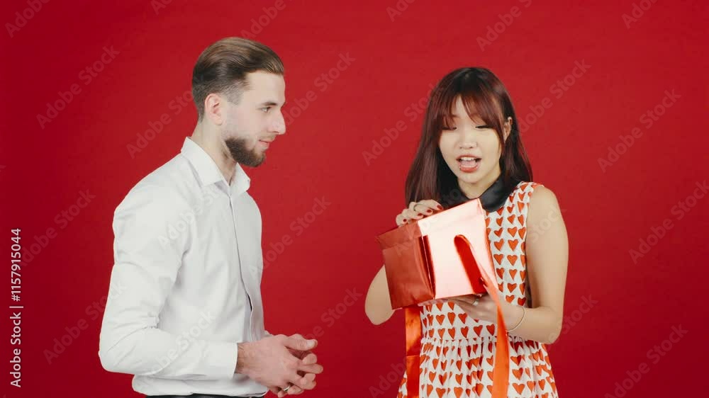 An Asian woman opens her Valentines Day gift from her boyfriend on a red background, but her disappointed expression shows it didnt meet her expectations. An Asian woman opens her Valentines Day gift from her boyfriend on a red background, but her disappointed expression shows it didnt meet her expectations.