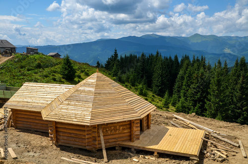 Rustic wooden cabin nestled in a lush, green mountainous landscape under a bright blue sky with fluffy clouds. Ideal for concepts of nature, tranquility, and rural living. Carpathians, Ukraine
 photo