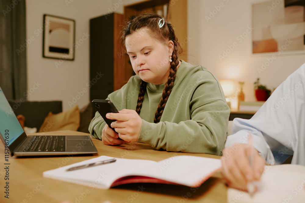 Medium shot of Caucasian girl with Down syndrome sitting at desk in living room, checking message on smartphone, hand of unrecognizable woman writing in notebook Medium shot of Caucasian girl with Down syndrome sitting at desk in living room, checking message on smartphone, hand of unrecognizable woman writing in notebook