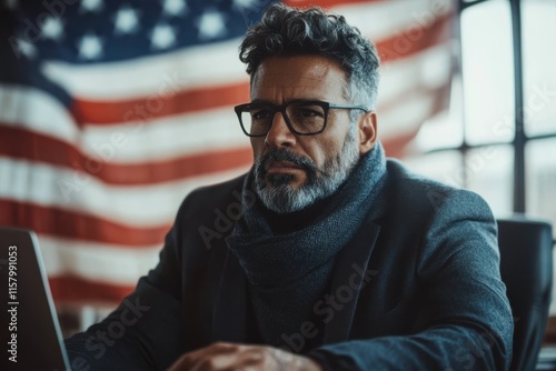 A man with an intense expression sits before a laptop in a warmly lit room, wearing glasses and a scarf, while an American flag proudly hangs in the background. photo