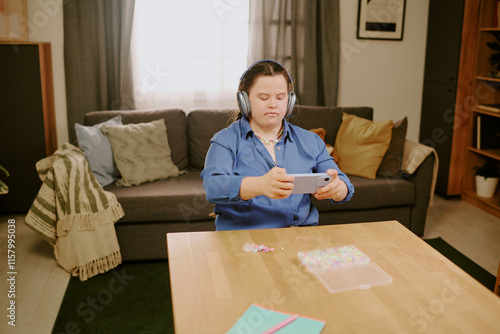 Creative girl with Down syndrome wearing headphones sitting at desk and taking photos of work in progress on smartphone, while beading photo