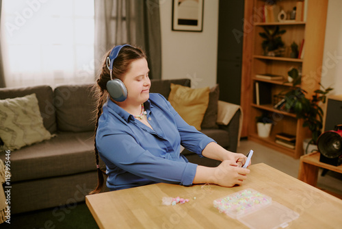 Cheerful girl with Down syndrome wearing headphones setting front-facing camera on smartphone for selfie, while sitting at desk and beading photo