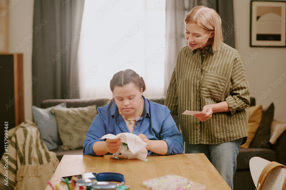 Obraz premium Mature Caucasian woman standing at desk in living room, watching her daughter with Down syndrome embroidering