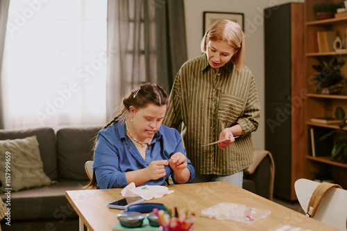 Loving mother standing at desk with pattern image in hand, helping her daughter with Down syndrome with embroidery process photo