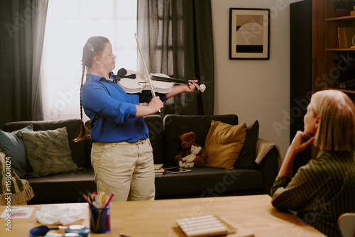 Modern girl with Down syndrome standing in living room, playing violin in front of her proud mother photo