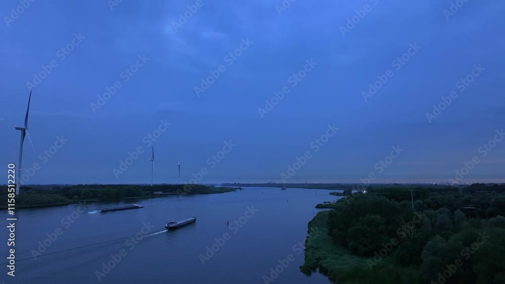 Aerial overview of a large barge transporting containers during the blue hour, gliding on calm waters below wind turbines Aerial overview of a large barge transporting containers during the blue hour, gliding on calm waters below wind turbines