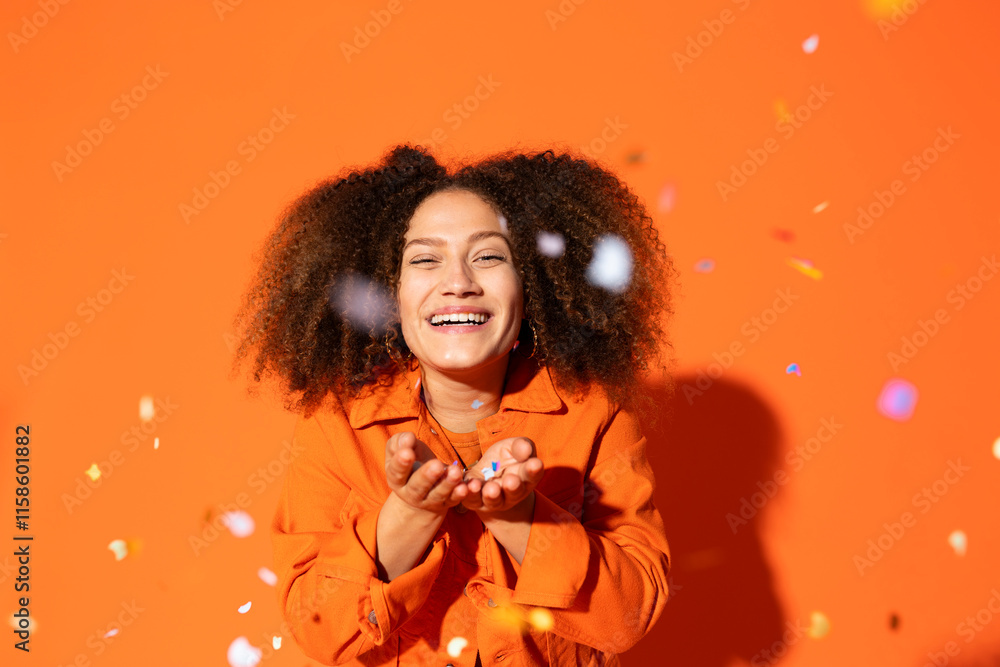 Smiling woman blowing confetti in front of orange background Smiling woman blowing confetti in front of orange background