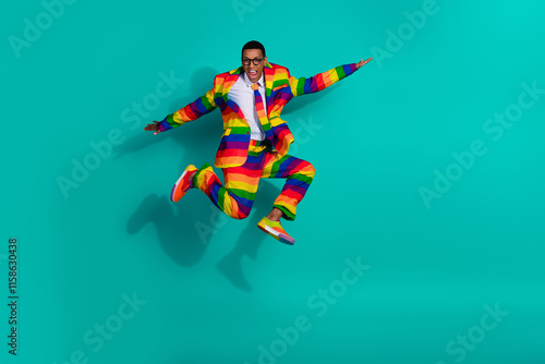 Charismatic man in vibrant rainbow-striped suit enthusiastically jumping against bright turquoise background photo