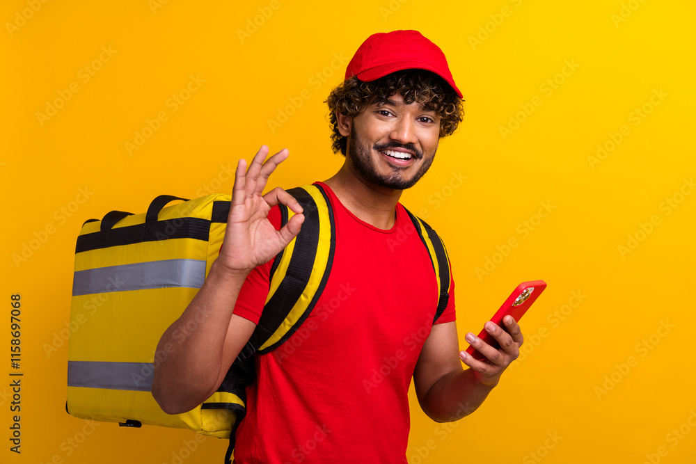 Young delivery worker holding a smartphone and smiling, wearing a red uniform and showing an okay sign with a cheerful expression. Young delivery worker holding a smartphone and smiling, wearing a red uniform and showing an okay sign with a cheerful expression.