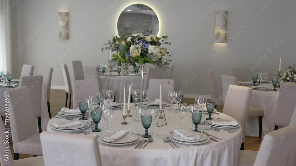 Elegant wedding reception table setting with white tablecloths, tall floral centerpieces of white and blue flowers, and neatly arranged glassware, candles, and dinnerware Elegant wedding reception table setting with white tablecloths, tall floral centerpieces of white and blue flowers, and neatly arranged glassware, candles, and dinnerware