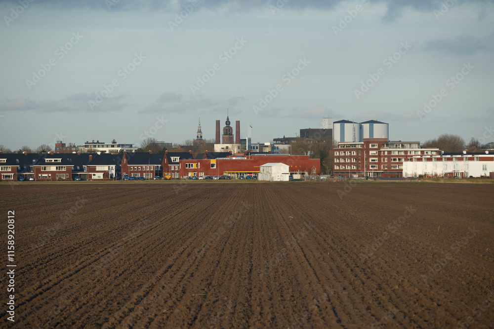 Plowed field foregrounds residential and industrial buildings in a small town.  Cloudy sky above.  A muted palette.