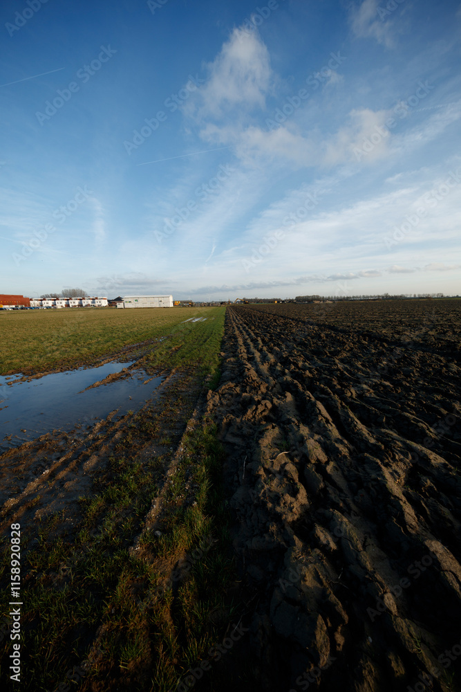 Plowed field next to grassy area with buildings. Sunny day with partly cloudy sky. Muddy tracks evident. Plowed field next to grassy area with buildings. Sunny day with partly cloudy sky. Muddy tracks evident.