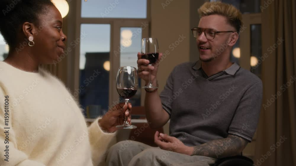 Tilt shot of young Black woman in cozy loungewear and young Caucasian man in wheelchair raising glasses of red wine in toast spending romantic relaxed evening at home together Tilt shot of young Black woman in cozy loungewear and young Caucasian man in wheelchair raising glasses of red wine in toast spending romantic relaxed evening at home together