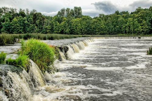 wide and low waterfall at Kuldiga city in Latvia. Water foam and forest in the background
