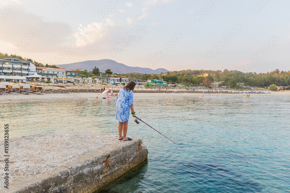 Naklejka premium Woman fishing on a pier, gazing over a serene beach resort, calm waters, and distant hills.