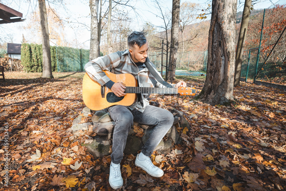 Focused guitarist in a plaid shirt strums an acoustic guitar while sitting on a stone bench amidst a golden autumn landscape. Sunlight enhances the warm, peaceful setting Focused guitarist in a plaid shirt strums an acoustic guitar while sitting on a stone bench amidst a golden autumn landscape. Sunlight enhances the warm, peaceful setting