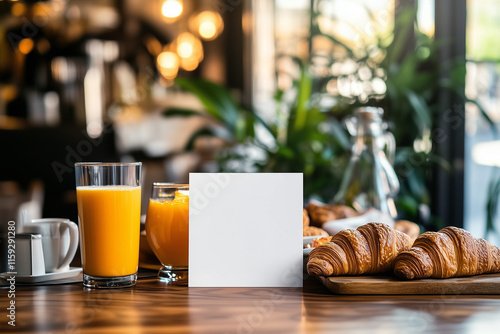 A blank white mockup greeting card is placed standing near a brunch spread with croissants, orange juice, and coffee in a casual restaurant.