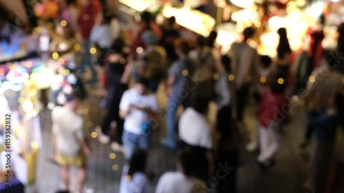 A crowd of people are walking down a street with a blurry background. Scene is lively and bustling, with people going about their day