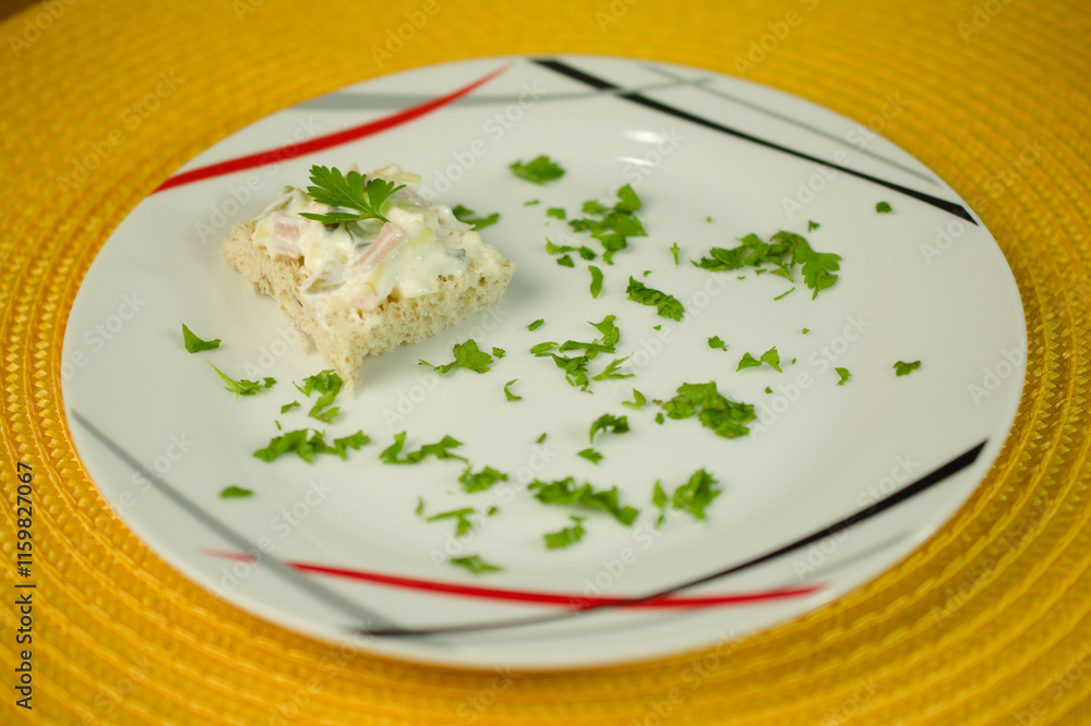 A single piece of whole wheat bread topped with egg salad and parsley, surrounded by scattered parsley on a modern-patterned plate with a vibrant yellow background. A single piece of whole wheat bread topped with egg salad and parsley, surrounded by scattered parsley on a modern-patterned plate with a vibrant yellow background.