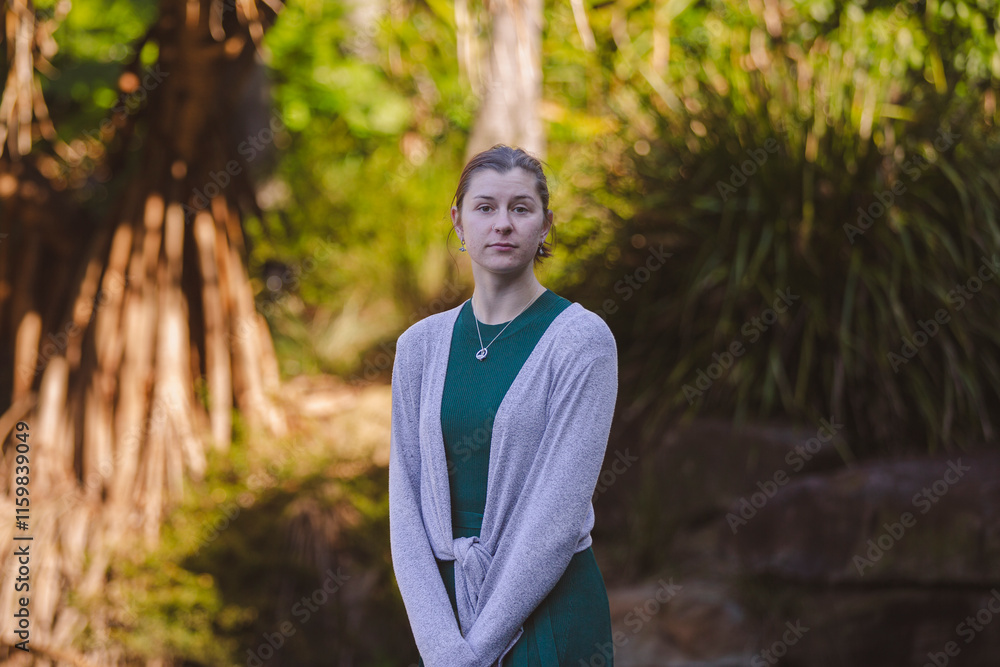 A smiling woman in a green dress with a gray cardigan stands outdoors, framed by lush green plants and soft sunlight, creating a serene, natural atmosphere A smiling woman in a green dress with a gray cardigan stands outdoors, framed by lush green plants and soft sunlight, creating a serene, natural atmosphere