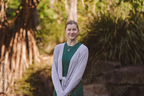 A smiling woman in a green dress with a gray cardigan stands outdoors, framed by lush green plants and soft sunlight, creating a serene, natural atmosphere