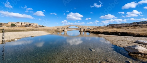 Ancient stone bridge reflecting in calm river under blue sky.