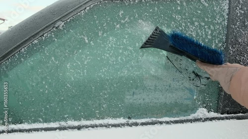 A person cleaning the side windows of a car from snow with ice scraper. Woman removes ice from car windows. Female hand cleans car with special tool in snowy winter day. 