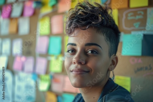 happy non binary person with short hair looking at camera near corkboard with paper notes in office photo