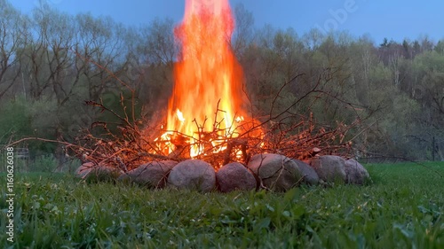 A large bonfire burning brightly in a grassy field. The fire is surrounded by a pile of branches and rocks and the grass, adding depth and context to the scene