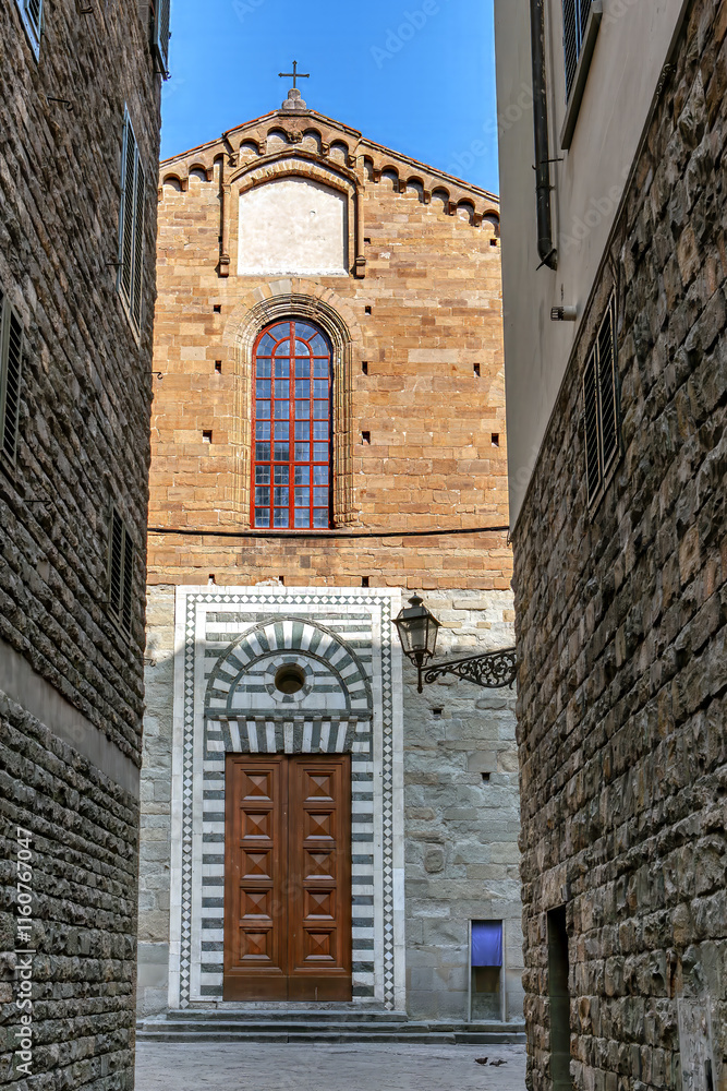A beautiful church with a large wooden door and intricate stonework stands between narrow alleys in Florence, Tuscany under a bright blue sky.
