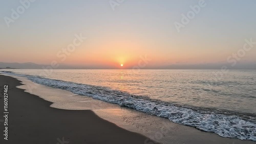 Handheld video of the sun at sunrise or sunset over the horizon of the calm Mediterranean Sea, with clear sky at golden hour, seen from the beach with the waves coming over the sand