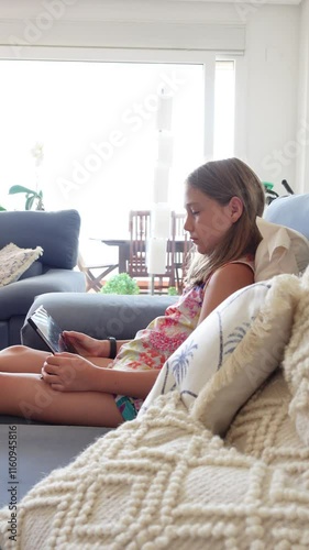 Vertical video of a pre teen girl looking at a tablet while sitting comfortably on the sofa at home
