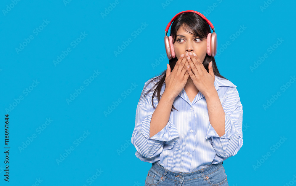 Young woman wearing pink headphones shows surprise against a bright blue background while covering her mouth Young woman wearing pink headphones shows surprise against a bright blue background while covering her mouth