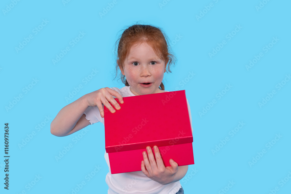 Excited child reveals bright red gift box against a vibrant blue background during a playful moment Excited child reveals bright red gift box against a vibrant blue background during a playful moment