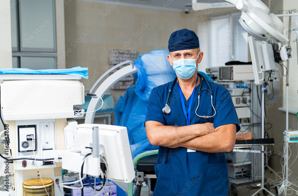 Naklejka premium Health pro in a modern hospital. A male doctor stands confidently in a hospital room, wearing scrubs and a mask, surrounded by medical equipment.