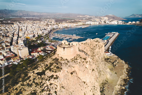 Aerial view castle of San Juan de las Aguilas, historic fortress perched on rocky hill overlooking town and Mediterranean Sea and marina with docked yachts and boats. Costa Calida, Murcia, Spain