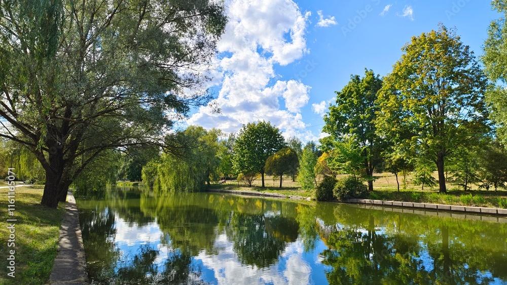 Obraz premium The banks of the river are concreted. Willows grow on the grass, their branches overhanging the water. There are ripples on the water. The trees and the sky with clouds are reflected in the water
