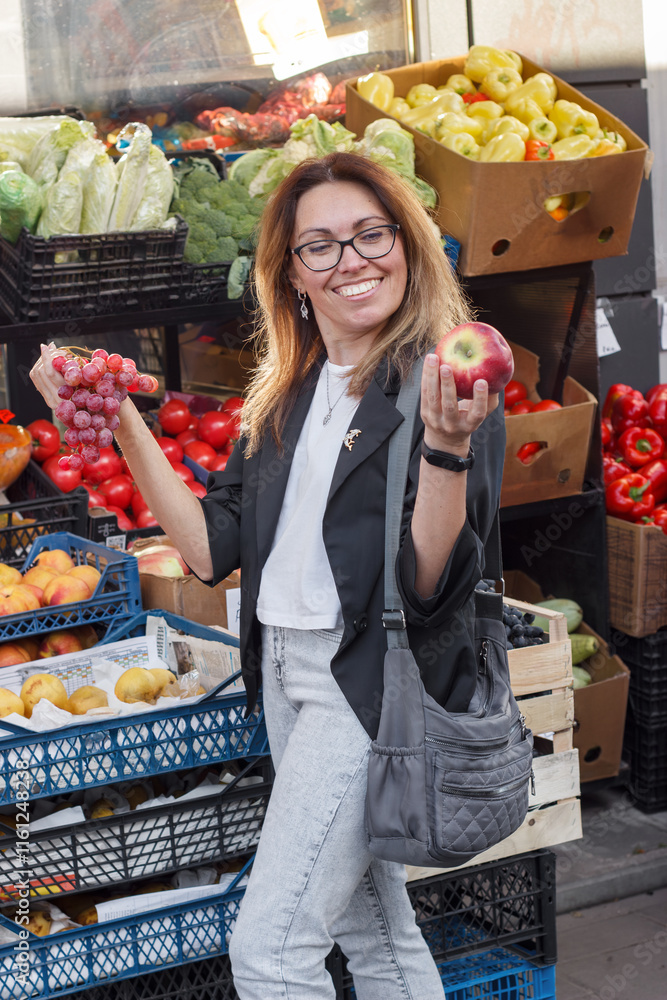 Obraz premium A woman in her 30s-40s selects fresh grapes at an outdoor market. The photo highlights organic produce, healthy eating, vegan lifestyle, sustainable living, and mindful nutrition choices