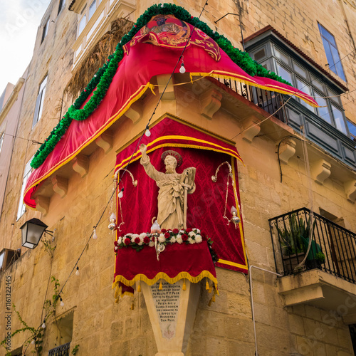 Valletta, Malta - February 9, 2024: Decorated Saint Paul statue on a pedestal on street corner in Valletta, ready for celebration with red and gold cloth and green garland