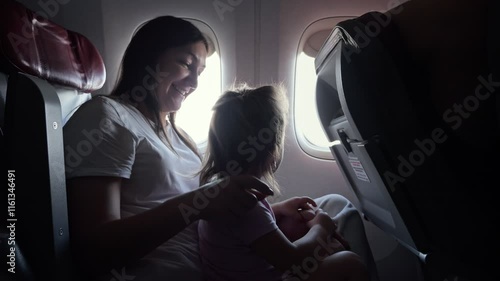 Happy woman plays with little daughter in cabin of airplane. Child admiringly follows scenery outside window hugging mother tenderly photo