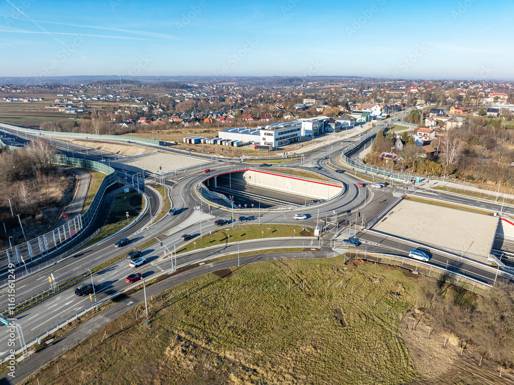 Fototapeta premium Krakow, Poland. Wegrzce interchange on new S52 highway (below) opened on December 23, 2024. Part of freeway around Cracow. Traffic circle with a hole inside and ramps. Aerial view in sunset light