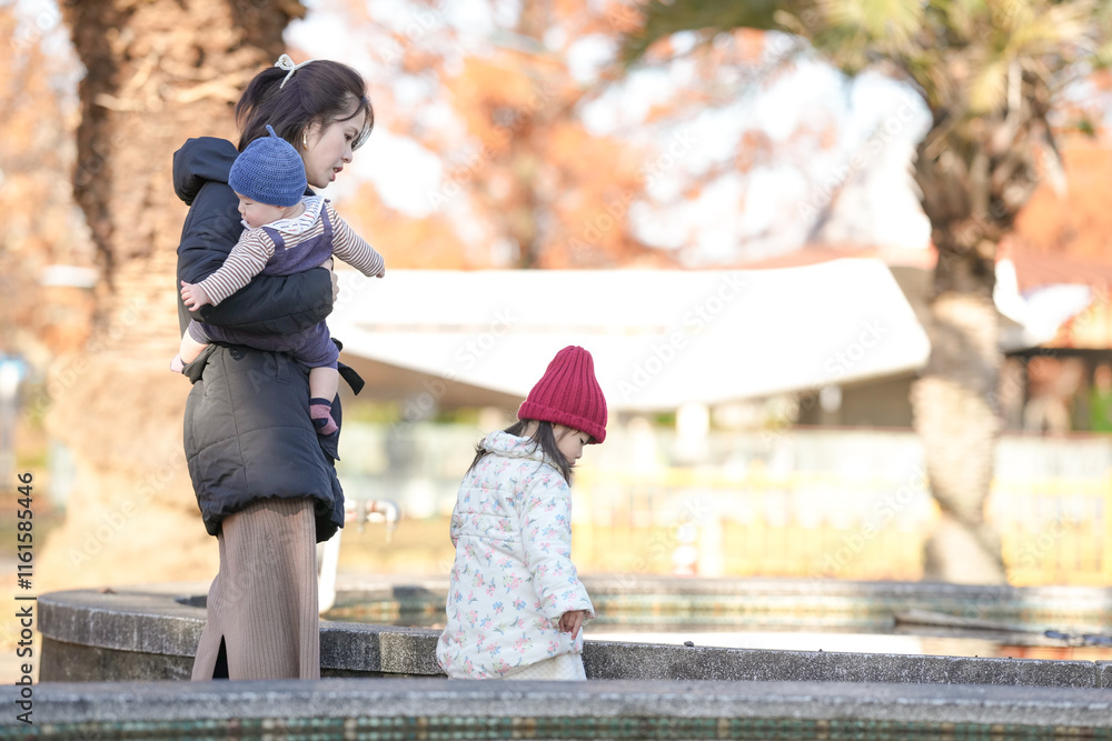 Obraz premium A 3-year-old Japanese girl wearing a down jacket, a 5-month-old baby, and a woman in her 30s walk by a pond in a park in Hakata City, Fukuoka Prefecture on a cold winter afternoon.