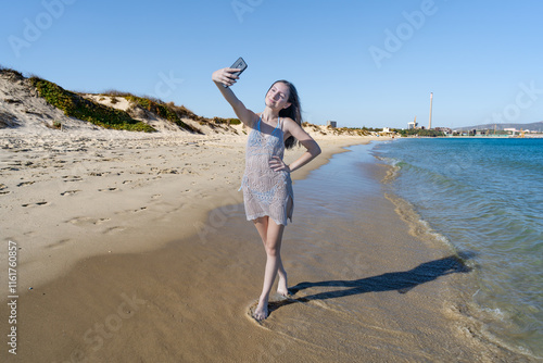 Chica joven en la playa con una sonrisa, haciendo selfi y videollamada mientras camina por la orilla sobre la arena un día de vacaciones en verano soleado y con cielo azul. photo