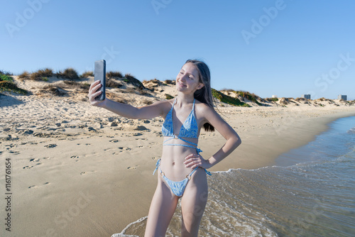 Chica joven en la playa con una sonrisa, haciendo selfi y videollamada mientras camina por la orilla sobre la arena un día de vacaciones en verano soleado y con cielo azul. photo