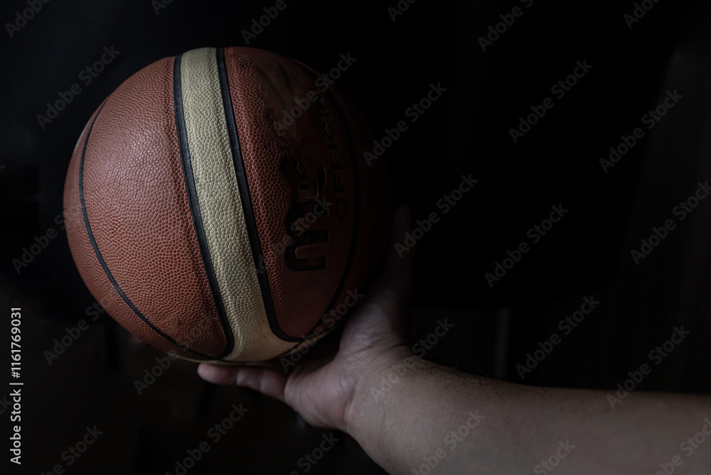 Silhouette view of Human hand holding basketball ball with confidence in minimalist style on black background. Close-up of A basketball leather in hand of a man against dark background, Light and Shad