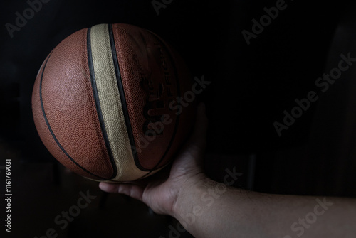Obraz na płótnie Silhouette view of Human hand holding basketball ball with confidence in minimalist style on black background. Close-up of A basketball leather in hand of a man against dark background, Light and Shad w salonie