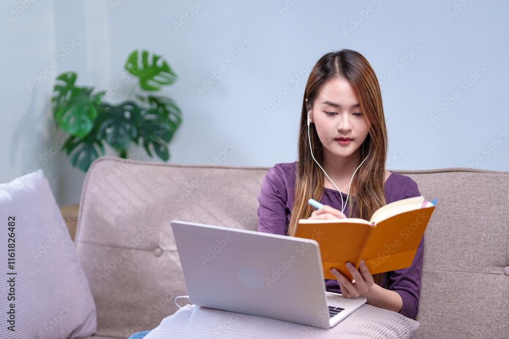 A woman sitting on a sofa with earphones, taking notes while attending an online class, using a laptop and a notebook A woman sitting on a sofa with earphones, taking notes while attending an online class, using a laptop and a notebook