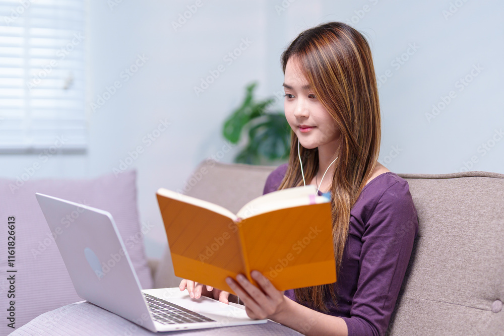 A woman sitting on a sofa with earphones, taking notes while attending an online class, using a laptop and a notebook A woman sitting on a sofa with earphones, taking notes while attending an online class, using a laptop and a notebook